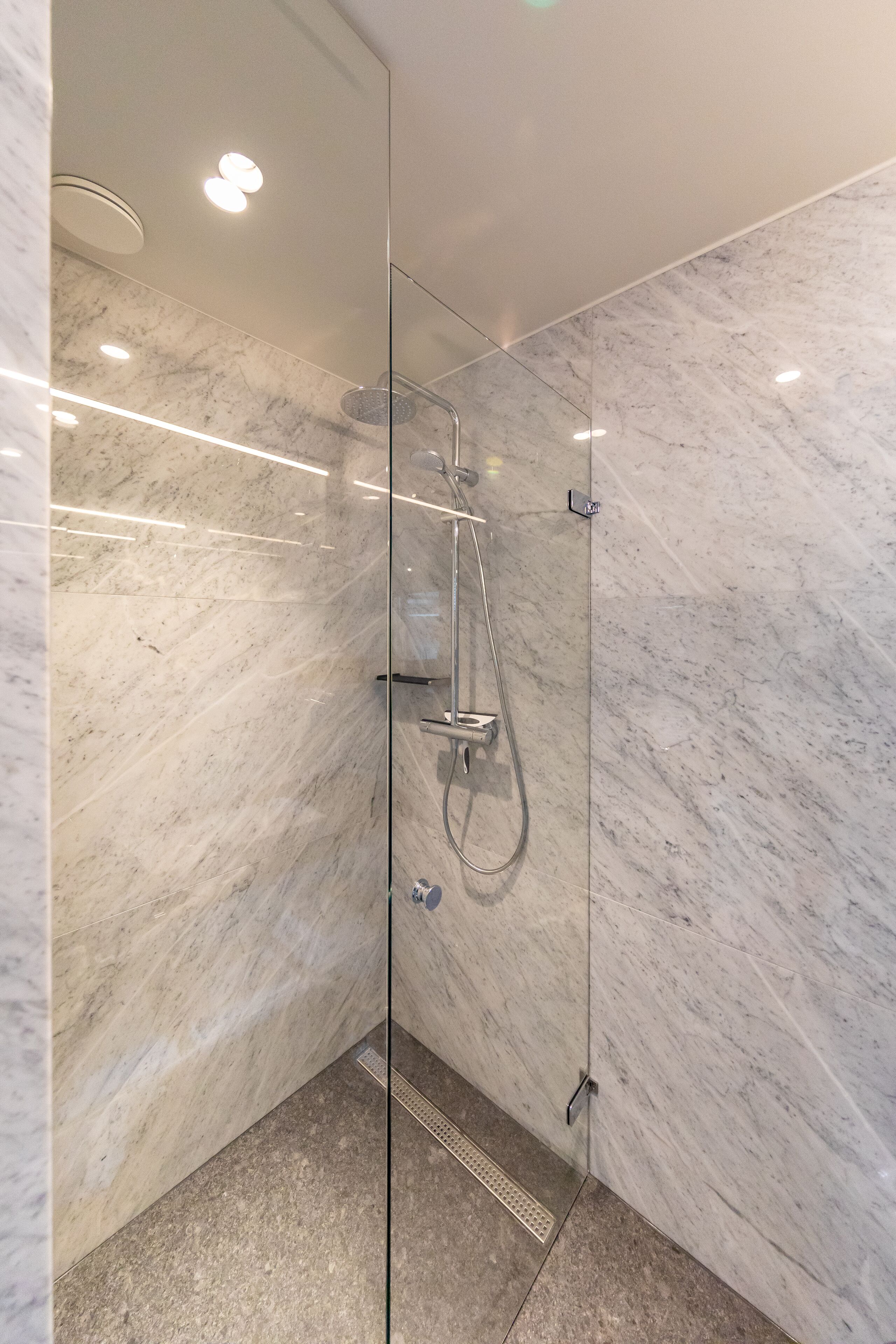 Un baño moderno con ducha de vidrio, paredes de mármol e iluminación empotrada en el techo.