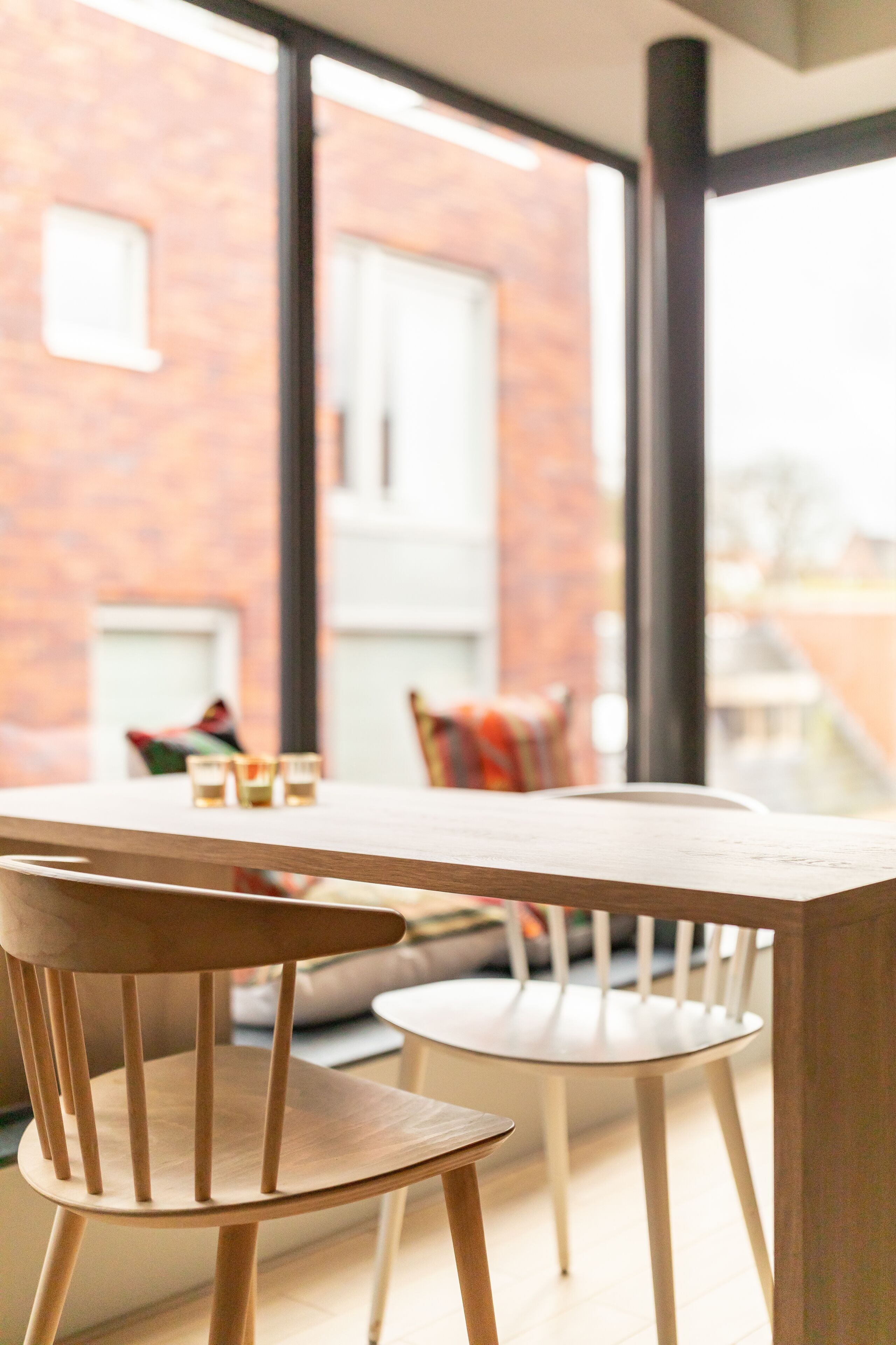 A modern dining area with a wooden table and chairs, large windows, and a brick wall in the background.