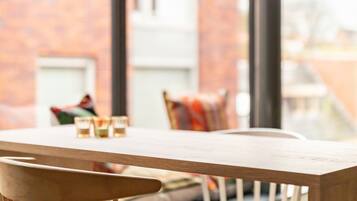 A modern dining area with a wooden table and chairs, large windows, and a brick wall in the background.