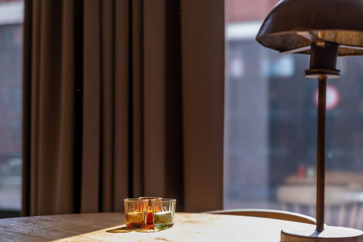 A table with a lamp and glasses on it, near a window with curtains.