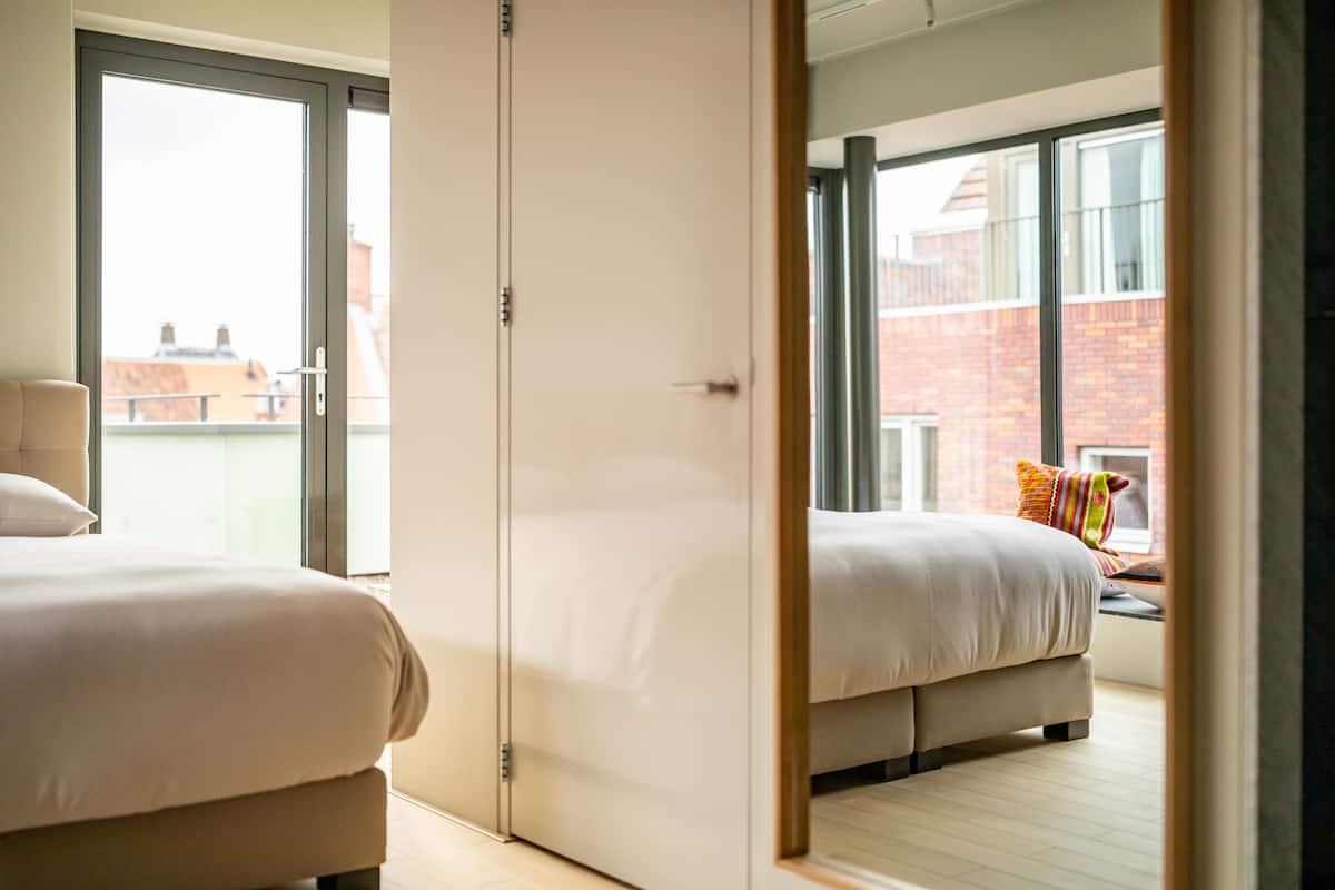 A modern hotel room with a large sliding door leading to a balcony, a bed with a white duvet, and a view of the cityscape.