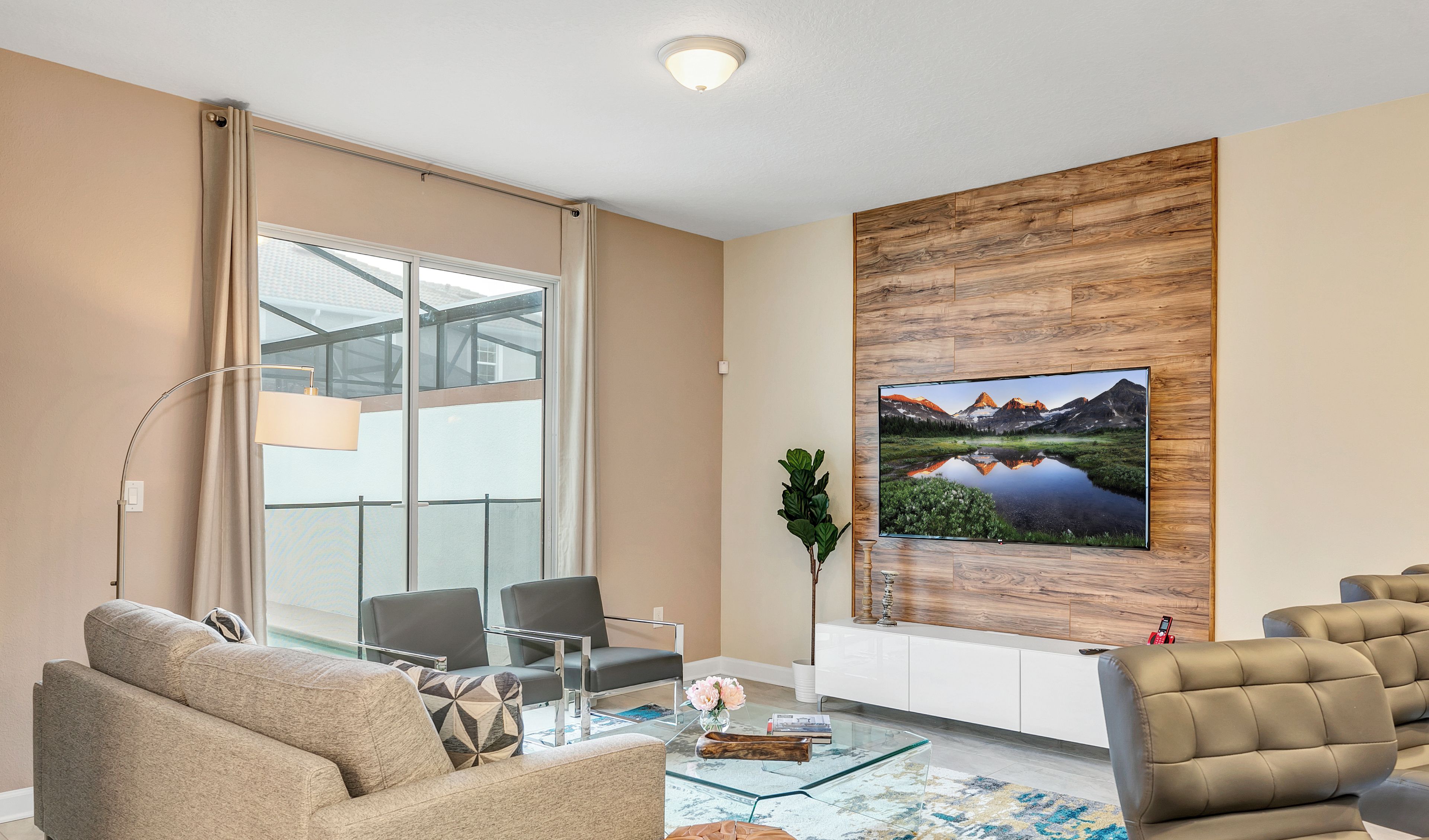 Een moderne woonkamer met een glazen salontafel, beige banken, een glazen eettafel en een grote flatscreen-tv bevestigd aan een houten wandpaneel.