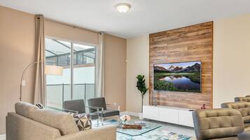 Een moderne woonkamer met een glazen salontafel, beige banken, een glazen eettafel en een grote flatscreen-tv bevestigd aan een houten wandpaneel.