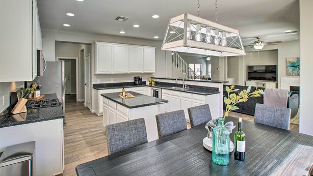 A modern kitchen with white cabinets, a central island, and a dining area with a dark wood table and grey upholstered chairs. - Game Room, Putting Green! Queen Creek Family Home (Queen Creek)