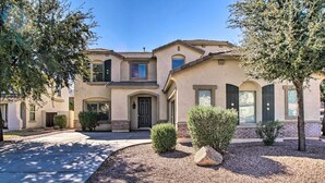 A two-story house with a beige facade, a front entrance with a door, and a driveway leading to the front door. There are two small bushes and a large rock in the front yard. - Game Room, Putting Green! Queen Creek Family Home (Queen Creek)