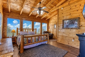 Une chambre dans une cabane en bois, avec un lit, un ventilateur de plafond, une cheminée et de grandes fenêtres.