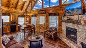 Interno di una baita di legno con zona pranzo, cucina, camino e ampie finestre.