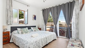 A bedroom with a large bed, a wooden nightstand, a bench, and a view of the outdoors through a sliding glass door. - Villa Caterina - Playa de Punta Prima (San Luis)