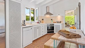 A modern kitchen with white cabinetry, a large window, and a dining area with a wooden table and wicker baskets. - Villa Sirena - Cap den Font (San Luis)