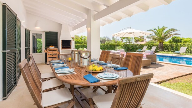 An outdoor dining area with a wooden table set for six, surrounded by wicker chairs, near a pool and greenery. - Villa Sirena - Cap den Font (San Luis)