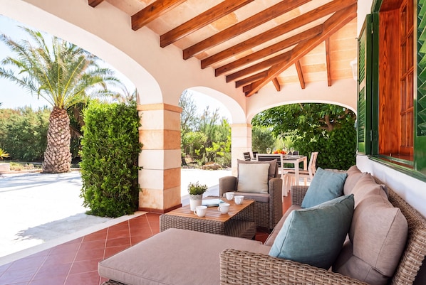 A covered outdoor seating area with wicker furniture, a table, and a view of the street and greenery.
