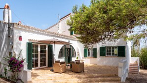 A white building with green shutters, a stone patio, and wicker furniture. - Hort des Milord - Villa en Es Castell (Es Castell)