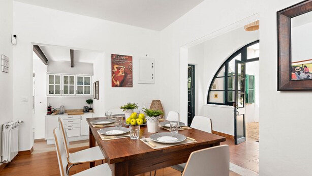A modern dining area with a wooden table set for four, white chairs, and a kitchen in the background. - Hort des Milord - Villa en Es Castell (Es Castell)