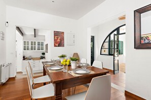 A modern dining area with a wooden table set for four, white chairs, and a kitchen in the background. - Hort des Milord - Villa en Es Castell (Es Castell)