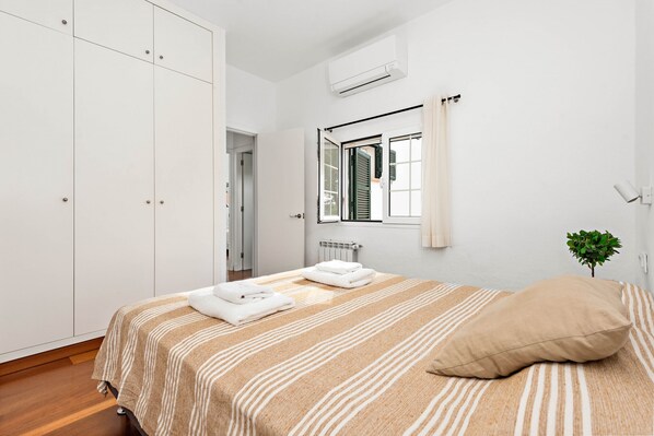 A bedroom with a bed, a wardrobe, an air conditioning unit, a window with a curtain, and a small plant. - Hort des Milord - Villa en Es Castell (Es Castell)