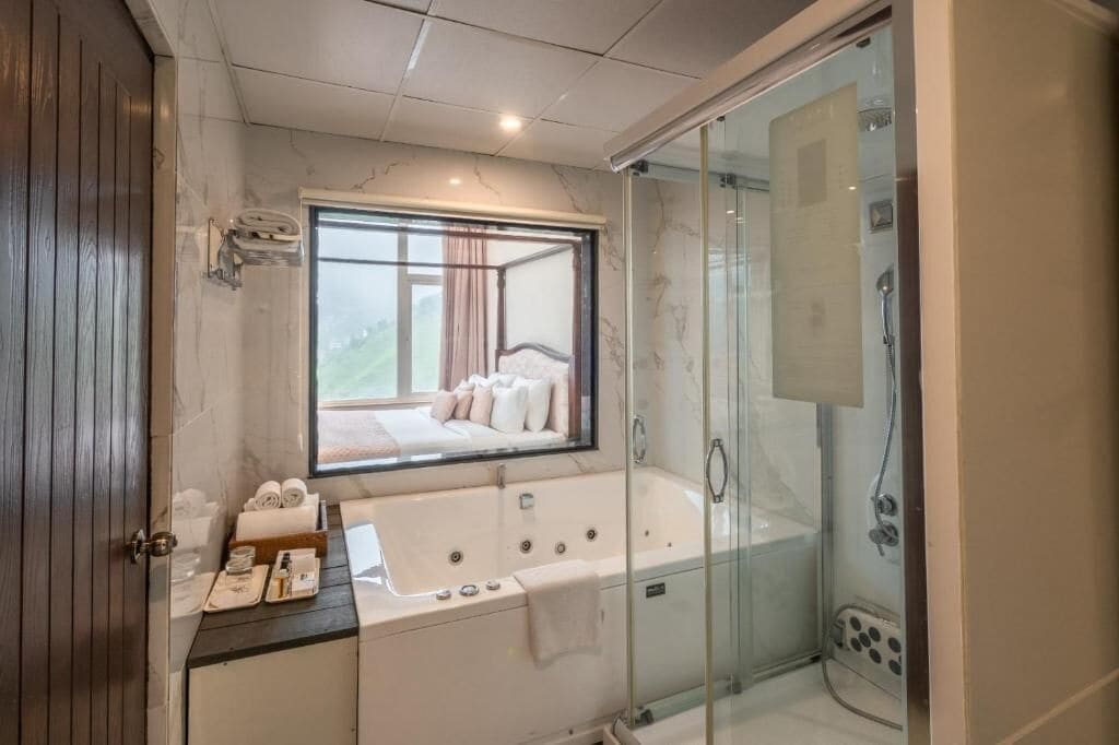 Un baño moderno con ducha de vidrio, una bañera grande y una ventana con vistas a un dormitorio.