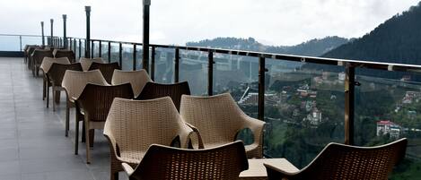 A row of wicker chairs and tables on a balcony with a glass railing, overlooking a mountainous landscape.