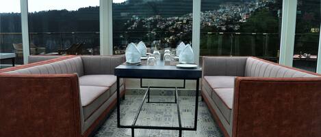 A dining area with a table set for four, two cushioned seating areas, and a view of a mountainous town through large windows.