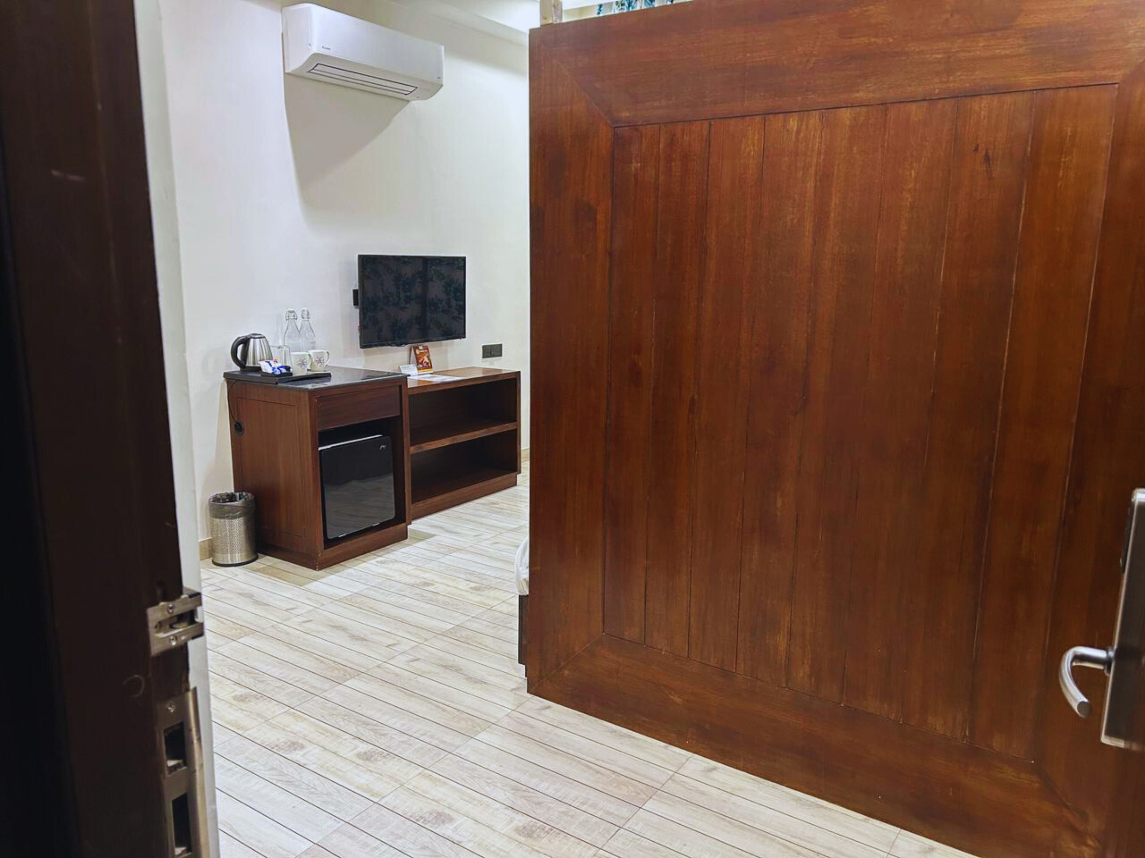 A room with a wooden door, a TV on the wall, a small cabinet, and a trash bin.