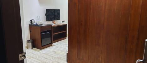 Una habitación con puerta de madera, televisor en la pared, un mueble pequeño y un cubo de basura.