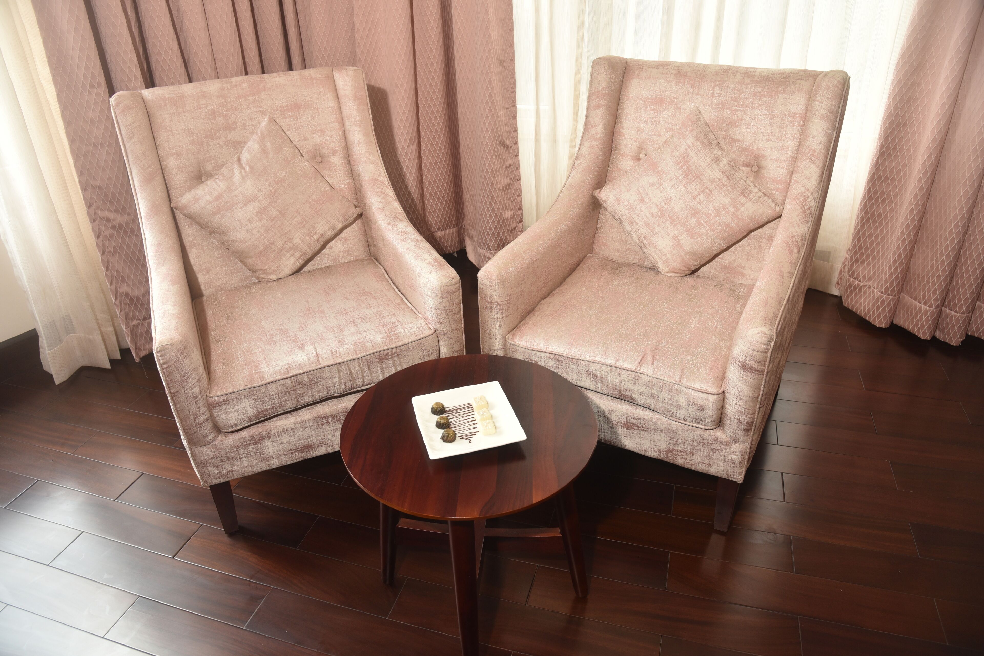 Two armchairs with a round wooden table between them, a plate with a decorative item, and a window with sheer curtains in the background.