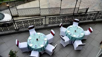 Two round tables with white chairs and a blue tablecloth, set on a tiled patio with a decorative railing in the background.