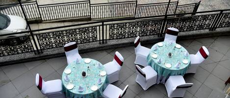 Two round tables with white chairs and a blue tablecloth, set on a tiled patio with a decorative railing in the background.