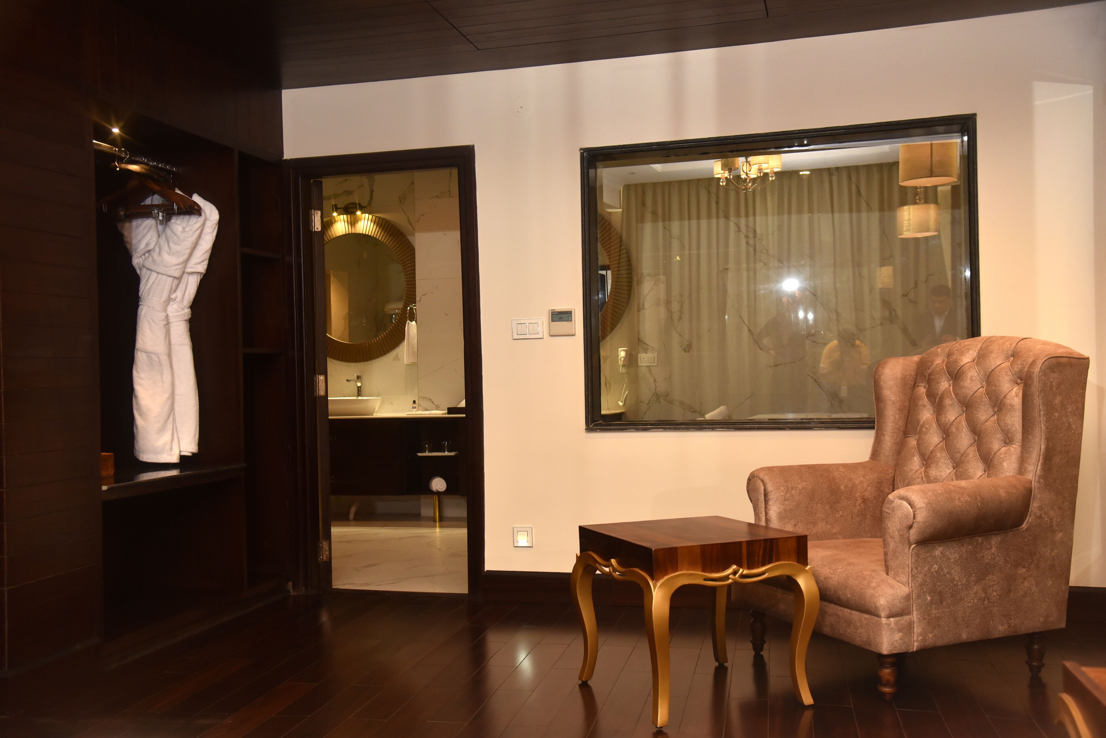 A room with a large mirror, a tufted armchair, a wooden table, and a robe hanging on a rack.