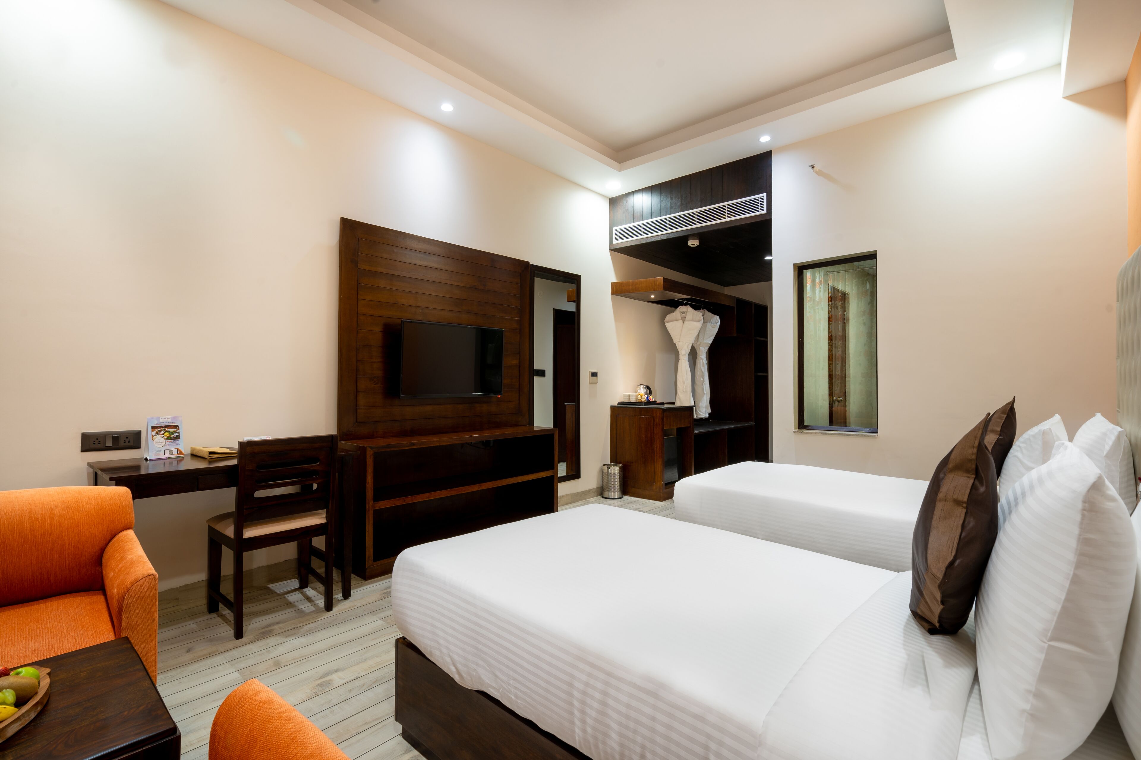 A hotel room with two beds, a desk, a chair, a TV, and a small table with a bowl of fruit.