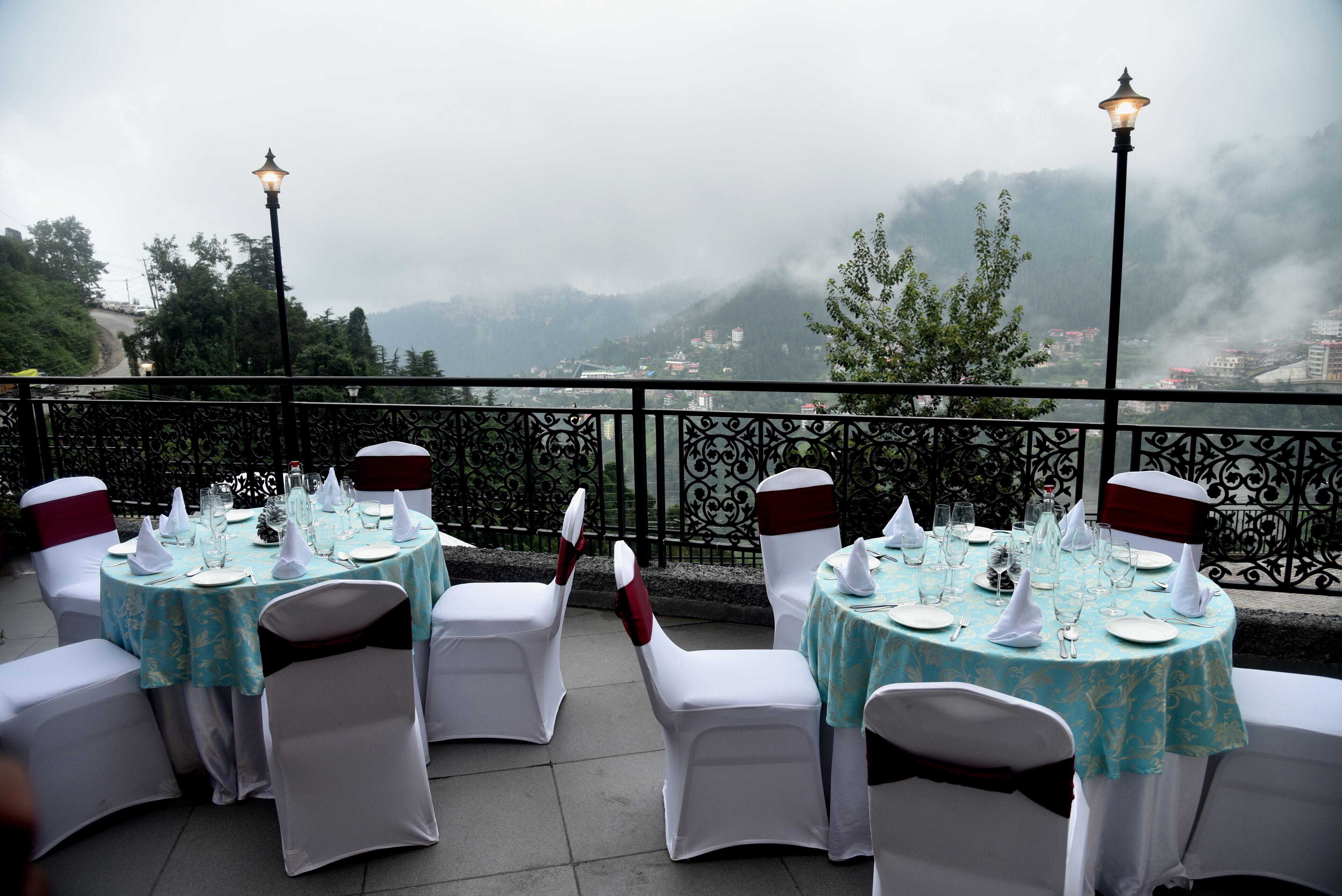 Una terraza con mesas redondas preparadas para comer, sillas blancas adornadas con cintas rojas, y vistas a montañas envueltas en niebla.