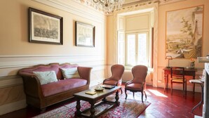 A room with a red leather sofa, two armchairs, a wooden coffee table, and a patterned rug. There are two framed pictures on the wall, a chandelier, and a large landscape painting. - La Corte. A historic house at the gates of Bergamo. (Mozzo - (Bg))