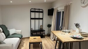 Una sala de estar acogedora con un sofá, una pequeña mesa de madera, un estante de metal negro y un comedor con mesa y sillas de madera.