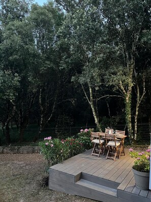 Una terraza de madera con una mesa y sillas, rodeada por un jardín con flores rosadas y una cerca.
