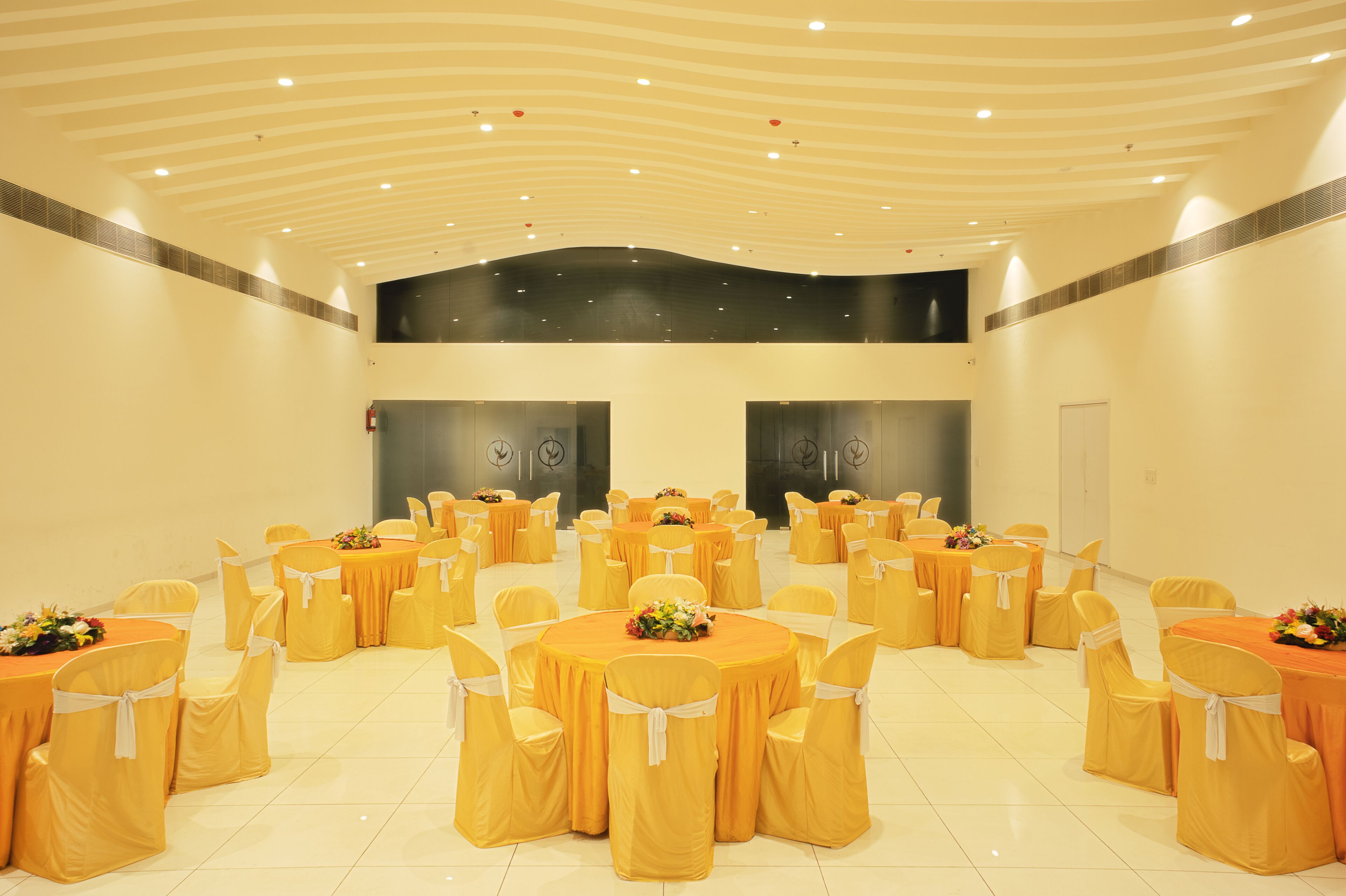 Une grande salle de conférence dotée de nombreuses tables rondes, chacune entourée de chaises recouvertes d’housses jaunes et agrémentées de rubans blancs. La salle est caractérisée par un plafond haut, un éclairage encastré et un design moderne.