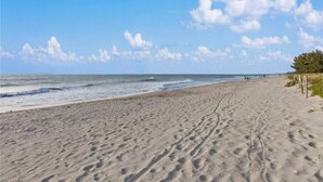 Una playa arenosa con huellas que conducen al agua, cielo despejado y una costa distante.