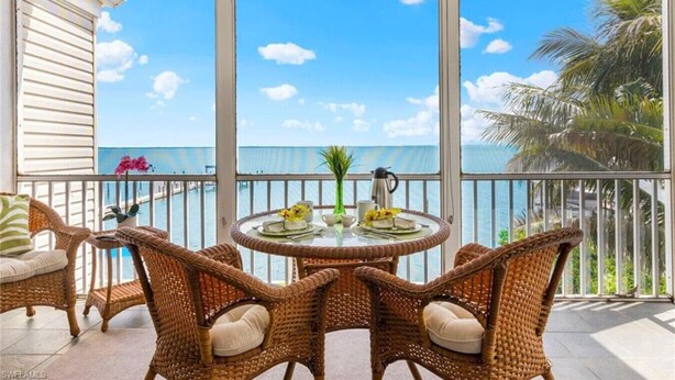 Un espacio de estar al aire libre techado con mobiliario de mimbre y una mesa preparada para comer, con vistas a un cielo azul despejado y un cuerpo de agua.