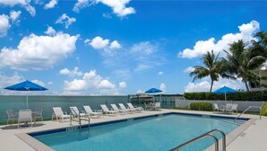 A swimming pool with lounge chairs and umbrellas, a clear sky, and a view of the ocean. - Captiva Hide-A-Way 1A (Captiva)