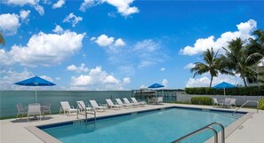 A swimming pool with lounge chairs and umbrellas, a clear sky, and a view of the ocean. - Captiva Hide-A-Way 1A (Captiva)