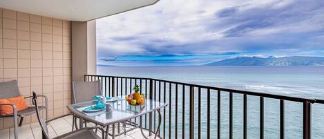 Een balkon met een tafel gedekt voor twee personen, een stoel en een schaal met fruit. Het balkon kijkt uit op een wateroppervlak met een bergketen in de verte.