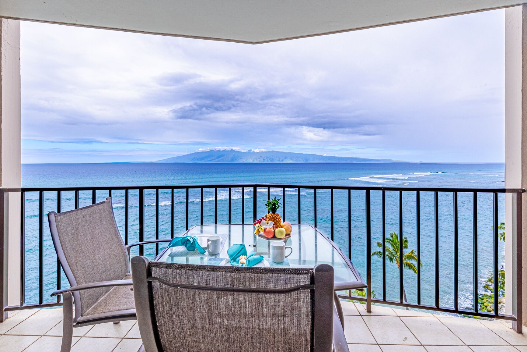 A balcony with a glass table and chairs, overlooking a scenic ocean view with a mountain in the distance.