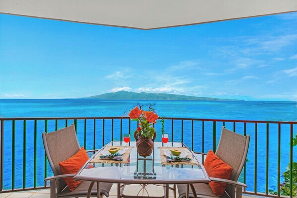 A balcony with a table set for two, overlooking a clear blue sea and a distant mountain.