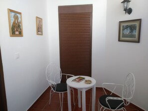 Una habitación pequeña con dos sillas de metal blanco, una mesita blanca con un libro y un objeto decorativo, y dos cuadros en las paredes.