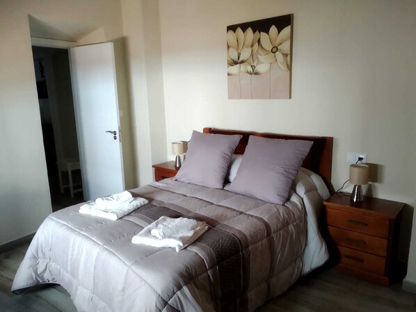 Una cama bien hecha con colcha color beige y marrón, dos mesitas de noche con lámparas y un cuadro de flores en la pared.