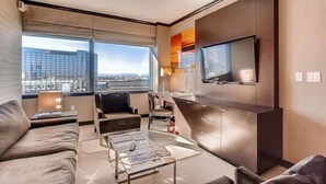 A modern hotel room with a large window offering a city view, a flat-screen TV mounted on the wall, a sofa, a coffee table, and a small dining area. - Perfect! Lux 1BR Suite at Vdara-15 (Las Vegas)