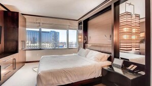 A modern hotel bedroom with a large bed, a desk with a phone, and a view of the cityscape through large windows. - Perfect! Lux 1BR Suite at Vdara-15 (Las Vegas)