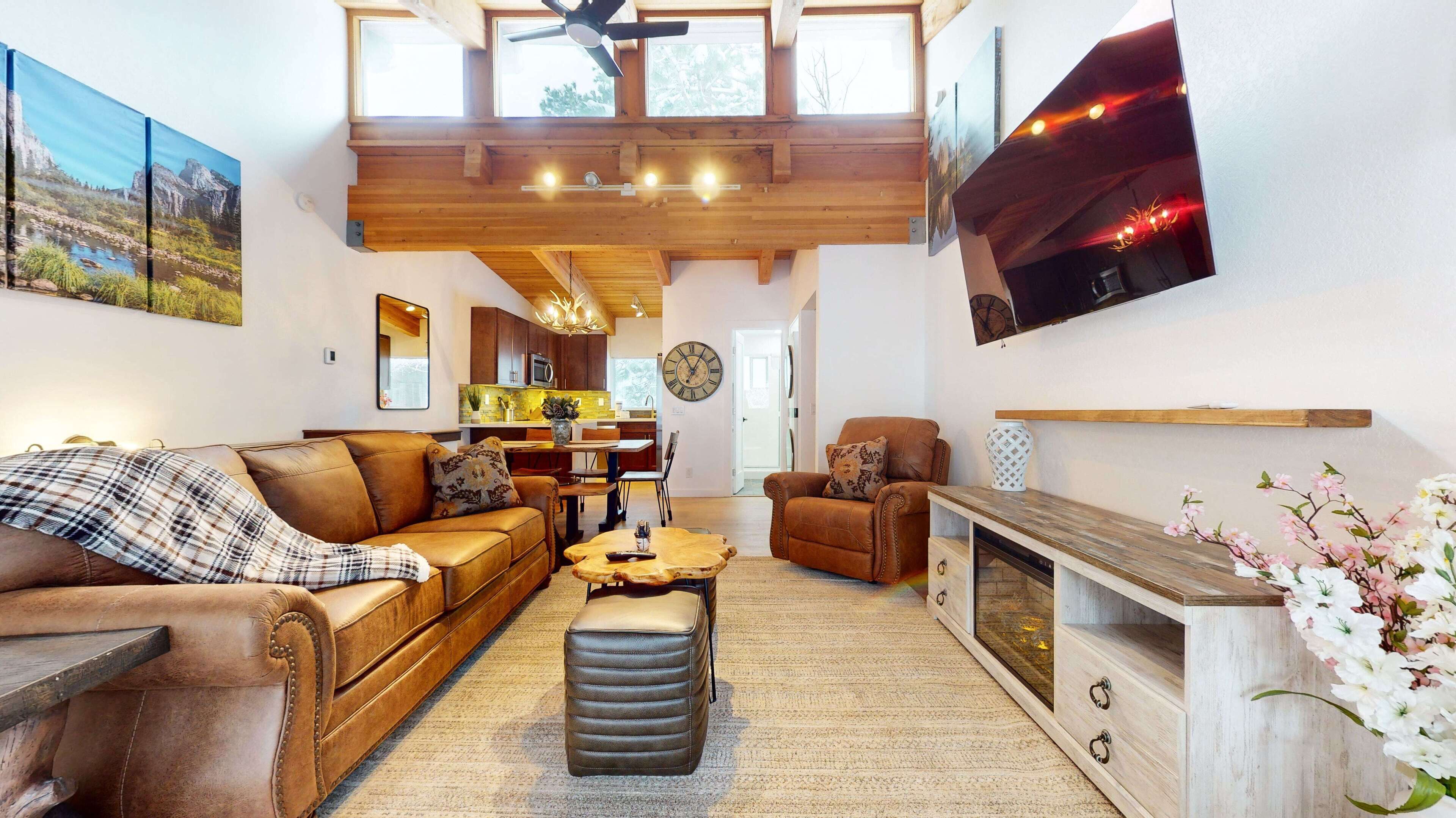 A spacious living room with a leather sofa, a coffee table, and a fireplace. There is a kitchen in the background with wooden cabinets and a dining area.