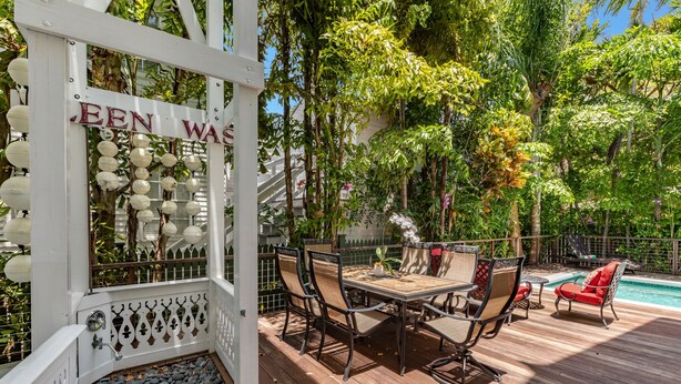 Eine Holzterrasse mit einer weißen Pergola mit der Aufschrift "JEEN WAS", umgeben von üppigem Grün und einem Pool.