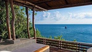 Une terrasse en bois avec des sièges extérieurs, une vue sur l’océan et un bateau visible au loin.