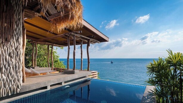Une piscine à débordement offrant une vue imprenable sur l’océan, une terrasse en bois avec des chaises longues et une structure surmontée d’un toit de chaume.