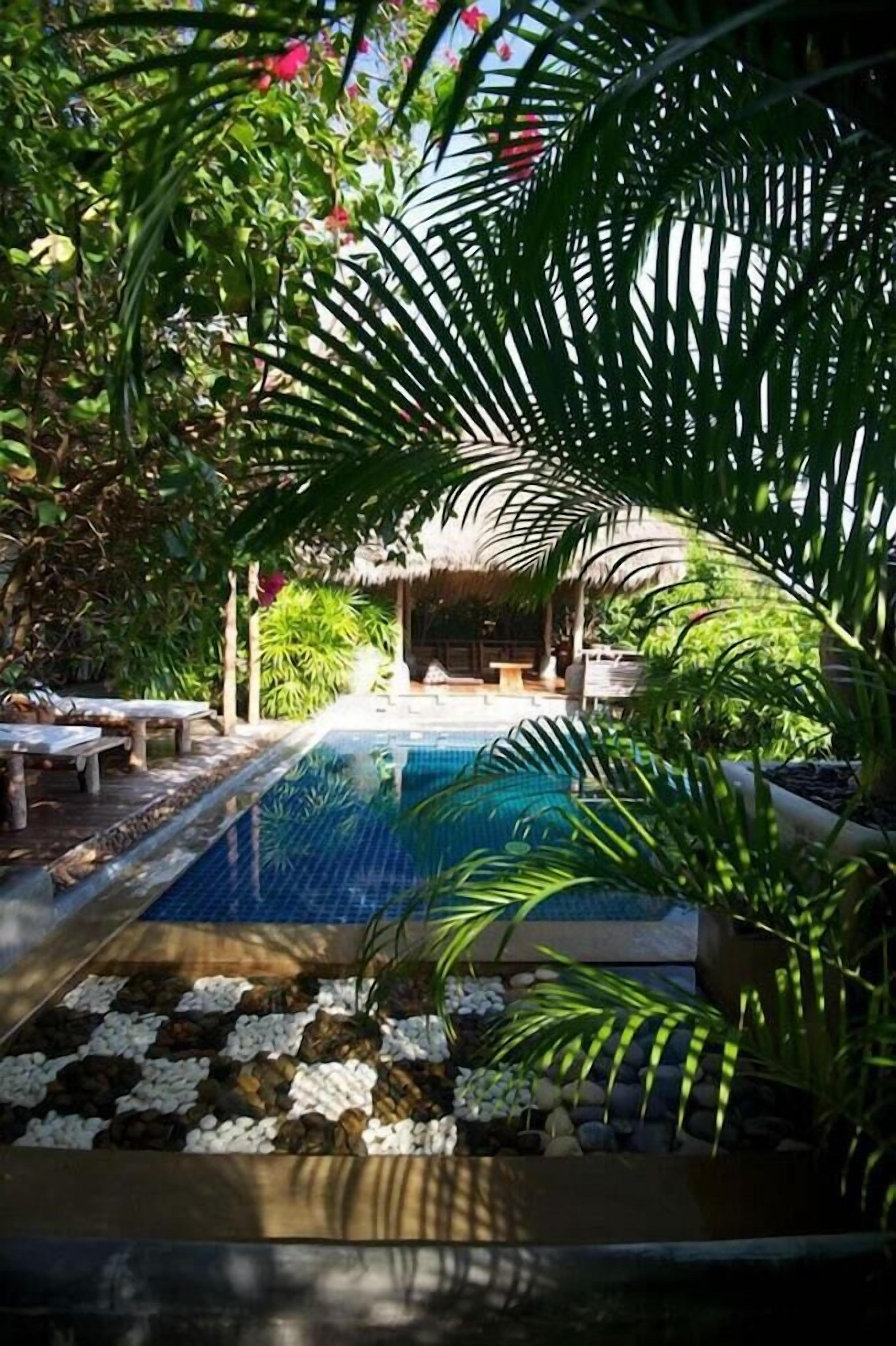 A pool area with a thatched roof structure, surrounded by lush greenery and a pebble-lined walkway.